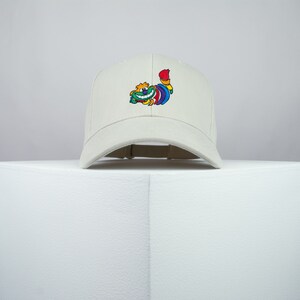 May include: A white baseball cap with a colorful cartoon character embroidered on the front. The character is a smiling creature with a rainbow tail.