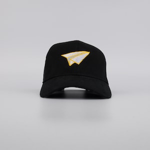 May include: Black baseball cap with a paper airplane design embroidered on the front. The paper airplane is white with yellow accents. The cap has a curved brim and a structured fit.