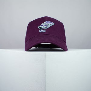 Puede incluir: Una gorra de béisbol de color morado con un diseño bordado de una consola de videojuegos y un mando. El texto "CONSOLE ME" está por encima de la consola.