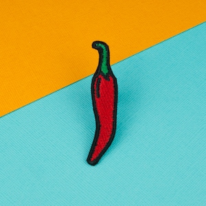 Puede incluir: Un parche de chile rojo bordado sobre un fondo de tela azul. El chile está bordeado de negro y tiene un tallo verde.