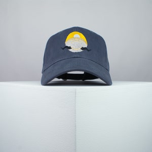 May include: A dark blue baseball cap with a yellow and white embroidered design. The design features a circle with a yellow top and a white bottom, with a black mustache underneath.