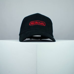 Può includere: Cappellino da baseball nero con logo Nintendo ricamato in rosso.