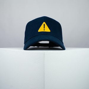 Op de afbeelding: Een donkerblauwe baseballpet met een gele driehoek en een uitroepteken.