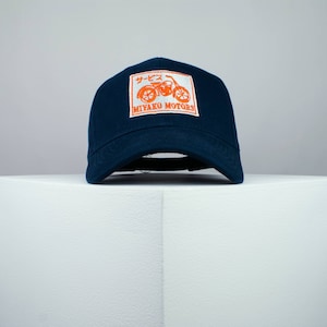 May include: A navy blue baseball cap with a white patch featuring an orange motorcycle and the text "サービス MIYAKO MOTORS"