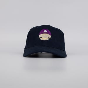 Puede incluir: Gorra de béisbol azul marino con un diseño bordado. El diseño presenta una forma morada y blanca con un contorno dorado. La gorra está hecha de una tela duradera y tiene una visera curva.
