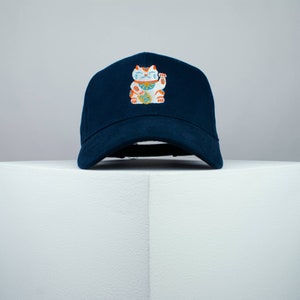 Op de afbeelding: Een donkerblauwe baseballpet met een geborduurd gelukkattenontwerp. De kat is wit met oranje en blauwe accenten en houdt zijn rechterpoot omhoog.
