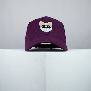 Può includere: Un berretto da baseball color borgogna con un cane di cartone animato con occhiali da sole ricamato sul davanti.