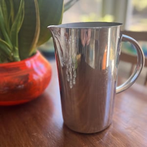 May include: A polished stainless steel pitcher with a cylindrical body, a curved handle on the side, and a small pouring spout at the top rim. The metal surface is reflective, showing light and surrounding shapes on its smooth exterior finish.