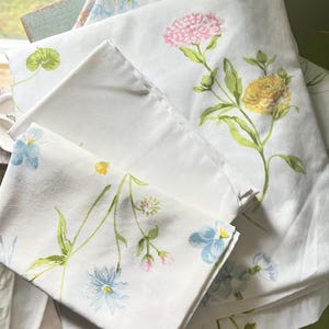 May include: A set of white bed sheets featuring a botanical print with various flowers in shades of pink, yellow, and blue. The fabric displays green stems and leaves scattered across the surface, with a folded pillowcase resting on top.
