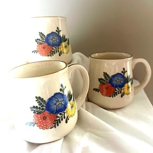 May include: Three ceramic mugs with floral designs. The mugs are white with a gold rim and feature a blue, yellow, and pink floral pattern.