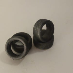 Può includere: Quattro piccoli oggetti neri a forma di anello con una superficie strutturata, forse pneumatici in miniatura o elementi decorativi. Gli oggetti sono disposti in gruppo, alcuni impilati e altri piatti.