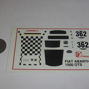 Peut inclure: Une planche de décalcomanies rectangulaire pour voiture miniature Fiat Abarth 1000 OTS, avec un motif à damier noir et blanc, le numéro 362, diverses formes noires, et le texte FIAT ABARTH 1000 OTS et B FIRENZE, présentée à côté d'une pièce.