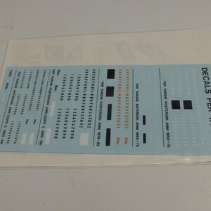 May include: A sheet of decals for number plates, with different fonts and numbers. The text includes "DECALS PER TARGHE" and several lines of numbers and letters. The decals are in black and white on a light blue background.