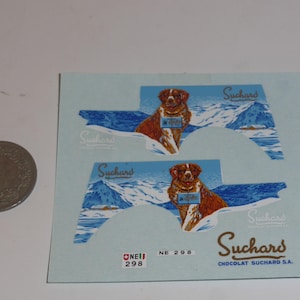 May include: Two Suchars chocolate stickers featuring a brown and white dog in a snowy landscape. The stickers have a light blue background with the Suchars logo and text. A coin is next to the stickers.