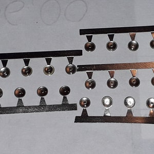 Può includere: Quattro strisce di metallo con piccoli cerchi rotondi attaccati a ciascuna striscia. I cerchi sono argentati e hanno un centro rialzato. Le strisce sono argentate e hanno uno sfondo nero.