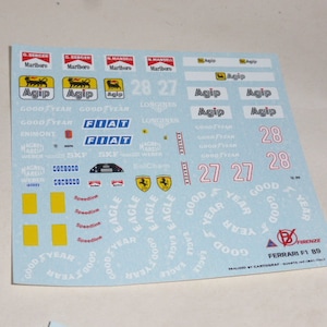 Puede incluir: Una hoja de calcomanías blanca con varios logotipos y números para un coche modelo Ferrari F1 89. Los logotipos incluyen Marlboro, Agip, Good Year, Fiat y Eagle. Los números incluyen 27, 28 y 28. La hoja también incluye el texto "FIRENZE" y "FERRARI F1 89".