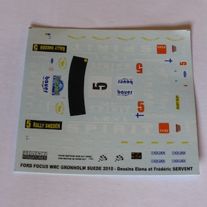 May include: Decal sheet for a model car, featuring the Ford Focus WRC Gronholm Suede 2010. The sheet includes various decals with the number 5, the name "bauer", and the text "Rally Sweden".