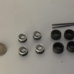 Könnte beinhalten: Ein Set aus vier silberfarbenen Miniatur-Mehrspeichenrädern mit schwarzen Reifen, zwei separaten schwarzen Gummireifen und zwei dünnen Metallachsen. Eine runde Münze liegt links daneben und dient als visueller Größenvergleich für die Teile.