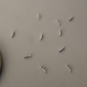 May include: A collection of small, thin, silver-colored metal pins or findings scattered across the frame, positioned next to a circular metallic coin that shows intricate embossed floral patterns on its surface for scale comparison.