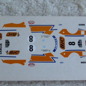 Puede incluir: Hoja de calcomanías con varias pegatinas para un coche modelo. Las pegatinas son de color naranja, azul y blanco con las palabras "Lancia", "Bic" y "Elbyn". El número "8" se repite en varias pegatinas.