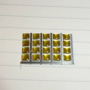 May include: A close-up of a gold-coloured circuit board with a grid of 24 small, square, yellow-tinted glass components.