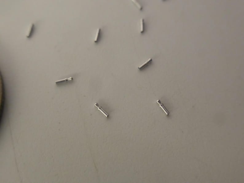 May include: A collection of small, silver-colored metal pins scattered across a surface. Each pin features a thin, cylindrical body with a slightly flared, rounded head at one end, resembling a miniature nail or a specialized jewelry finding component.