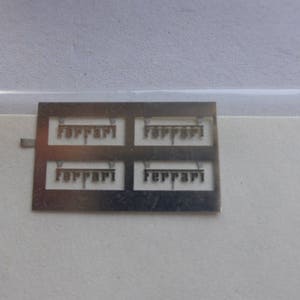 May include: A small, rectangular metal sheet with four cut-out "Ferrari" logos. The logos are in a serif font. The sheet is silver and sits on a white surface.