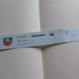 Peut inclure: Une feuille d'autocollants bleu clair avec divers logos et textes. Le logo "TAG Heuer" est en vert et rouge. D'autres logos incluent "Tecno Rally" et "Scuderia Vesuvio". Le texte "PRINTED BY CARTOGRAFIC.COM MADE IN ITALY" est également visible.