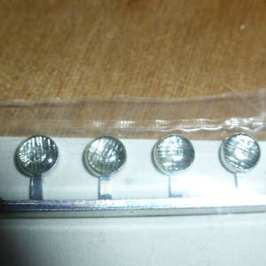 May include: Four miniature silver spotlights, each with a round, textured head, mounted on a silver bar. The bar is rectangular. The background is a light brown surface. The spotlights are likely for a model or miniature project.