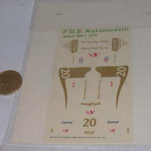 Puede incluir: Una hoja de calcomanías de carreras para un coche de carreras Wolf WR1-1978. Las calcomanías son principalmente doradas, blancas y rojas, con el número 20, el logotipo de Castrol y el logotipo de Goodyear. Una moneda de oro está en la imagen.