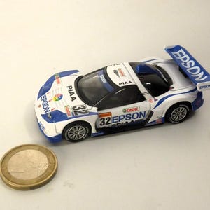 May include: A white and blue toy race car with the number 32 and the word "EPSON" printed on it. The car has a rear spoiler and is next to a gold coin.