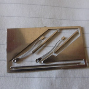 May include: A silver metal plate with a cut-out design of three overlapping lines. The lines are curved and have small circles at the ends.