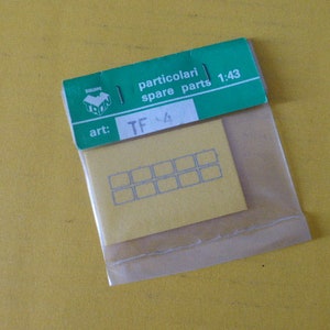 Op de afbeelding: Een kleine, doorzichtige plastic zak met een gele kaart met een raster van grijze rechthoeken en een groen label. Het label luidt "particolari spare parts 1:43" en "art: TF 4". De achtergrond is effen geel.