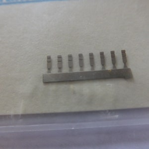 May include: A close-up of a silver strip with 12 small, rectangular metal pieces attached to it. The pieces are evenly spaced and appear to be part of a model kit.