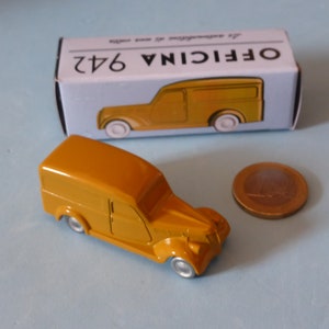 May include: A yellow toy van with a white roof and white wheels. The van is sitting on a blue surface. The van is next to a box with the words "OFFICINA 642" printed on it.