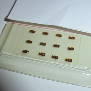 May include: A clear plastic package containing twelve small, rectangular, amber-colored gemstones. The gemstones are evenly spaced on a white card, with a red stripe visible on the package's edge. The package is partially open, revealing a white background.