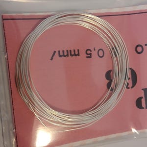 May include: A coil of thin, silver-colored wire, approximately 0.5 mm in diameter, is displayed. The wire is neatly coiled on a pink and red paper background, partially visible through a clear plastic bag.