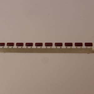 May include: A strip of ten small, rectangular, dark red components attached to a thin, gold-colored metal strip. The components are evenly spaced along the strip, which is set against a plain, light background.
