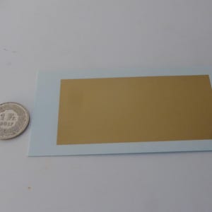 May include: A silver 1 Fr coin from 2017 next to a rectangular gold-colored sticker on a light blue card. The coin is on the left, and the sticker is centered on the card. The background is white.