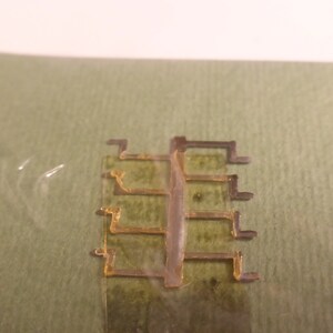 May include: A close-up of a gold-colored metal component with a clear plastic piece in the center. The component is on a green surface.