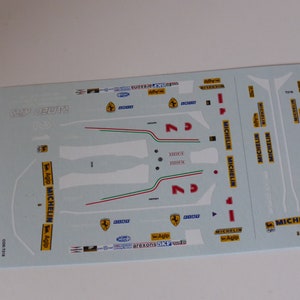 Puede incluir: Hoja de calcomanías con varias pegatinas para un coche modelo. La hoja presenta el logotipo de Ferrari, el número 2, las palabras "Agip" y "Michelin", y la bandera italiana.