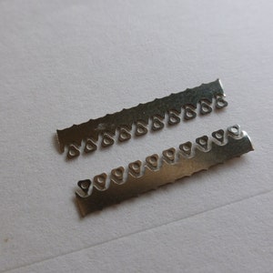 Könnte beinhalten: Zwei kleine, silberfarbene Metallstreifen mit dreieckigen Aussparungen an einer Kante. Die Streifen liegen auf einer weißen Oberfläche. Die Aussparungen sind gleichmäßig verteilt.