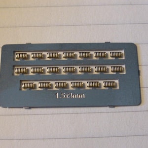 May include: A gray metal plate with 18 gold-colored rectangular components arranged in three rows of six. The plate has the text "1.5x3mm" printed on it.