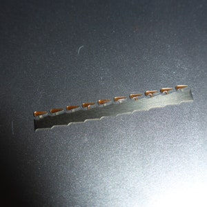 May include: A silver metal strip with a serrated edge and 15 small, brown, rectangular-shaped metal pieces attached to it.