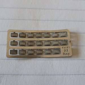 Könnte beinhalten: Ein silberner Metallstreifen mit 24 kleinen rechteckigen Löchern. Der Streifen trägt den Text "FARI 23 Shop".
