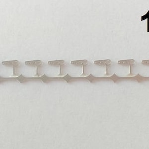 May include: A set of silver-colored metal garment hooks, arranged in a row. Each hook has a T-shaped top with three small holes and a diamond-shaped base. The text "1 set" is in the upper right corner.
