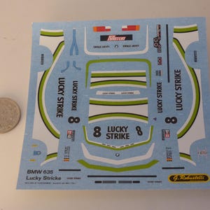 Puede incluir: Una hoja de calcomanías para un coche de carreras BMW 635, con el logotipo "Lucky Strike" y el número 8. Las calcomanías son principalmente azul claro, blanco y verde, con detalles en rojo y negro. Una moneda está al lado.
