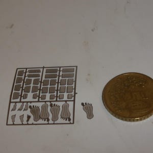 May include: A selection of small architectural model components and foot-shaped cutouts, possibly for a craft project, next to a gold-coloured coin. The tiny pieces are arranged on a white surface, suggesting a detailed miniature scene.