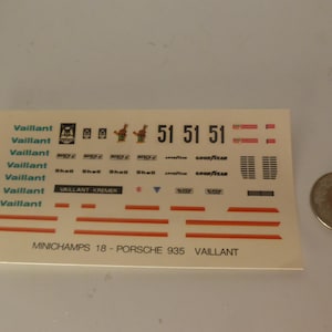May include: A sheet of decals for a model car, featuring the "Vaillant" logo and other graphics. The decals include the numbers "51" and various sponsor logos. A coin is next to the decal sheet.