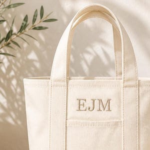 May include: Cream-colored canvas tote bag with a rectangular shape and two handles. The bag features a front pocket and embroidered initials "EJM". A seashell, books, and decorative beads are in the background.